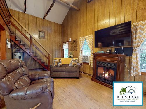 Cozy cabin interior with wooden walls, a fireplace, a sofa, and a recliner, featuring large windows and warm decor.