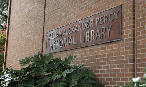 Sign for the William Alexander Percy Memorial Library on a brick wall, surrounded by green foliage.