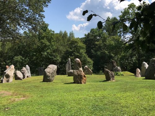 A circle of large standing stones in a grassy area, surrounded by trees and under a blue sky with clouds.