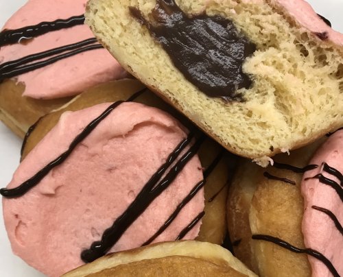 A close-up of fluffy donuts with pink frosting and chocolate drizzle, one cut open to reveal a chocolate filling.