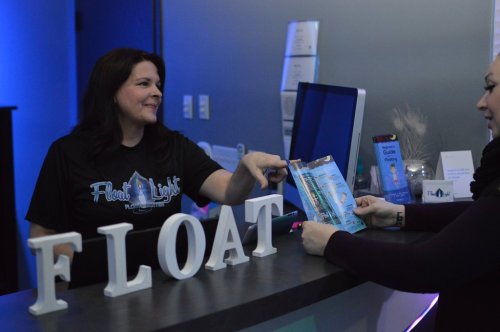 A woman at a reception desk hands a brochure to a customer in a softly lit environment. The word "FLOAT" is displayed prominently.