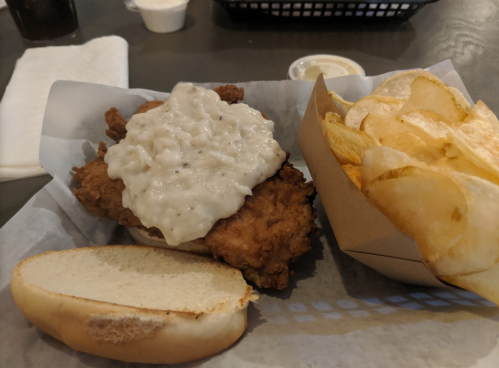A fried chicken sandwich topped with creamy gravy, served with a side of potato chips on a paper-lined tray.