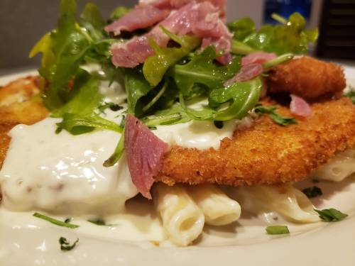 A plate of pasta topped with crispy chicken, creamy sauce, fresh greens, and slices of ham.