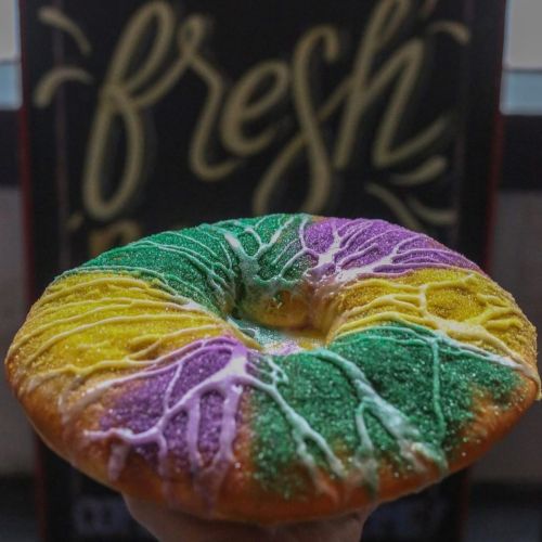 A colorful donut with purple, green, and yellow sugar, topped with white icing, held in front of a "fresh" sign.