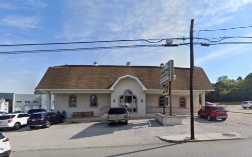 A large, light-colored building with a sloped roof, featuring multiple windows and a sign out front, surrounded by parked cars.