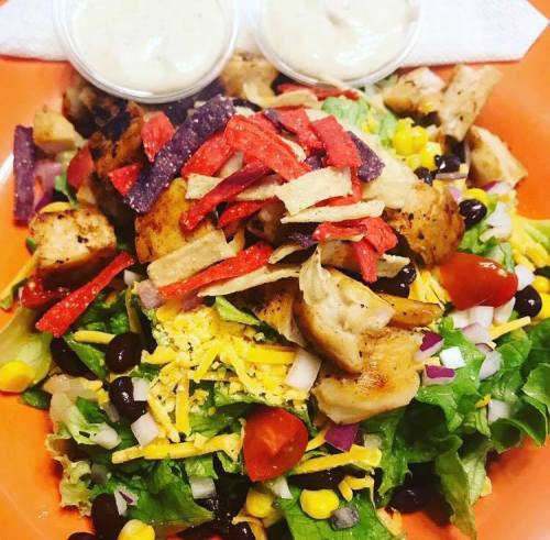 A colorful salad topped with grilled chicken, tortilla strips, cheese, and two cups of dressing on the side.