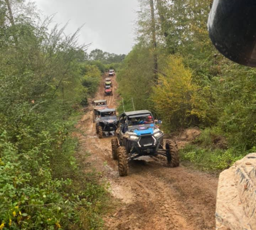 A line of off-road vehicles navigating a muddy trail surrounded by trees and greenery.
