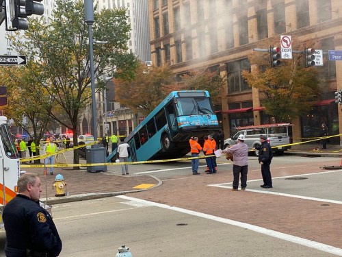 A bus is tipped over on its side in a city intersection, surrounded by emergency responders and caution tape.