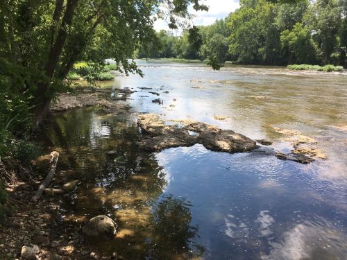 A serene river scene with rocky banks, lush greenery, and clear water reflecting the sky.