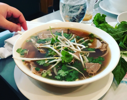 A bowl of pho with herbs, bean sprouts, and sliced meat, accompanied by a glass of water on a table.
