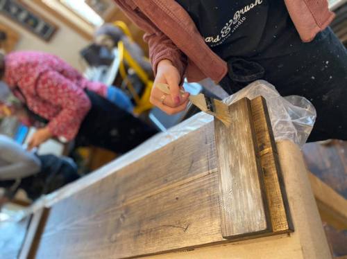 A person paints a wooden surface with a brush, while another person works in the background.