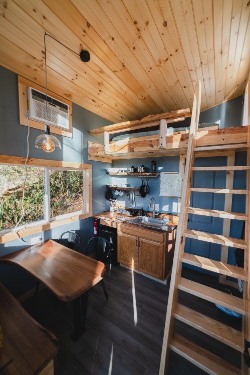 Cozy tiny house interior featuring a lofted bed, kitchen area, dining table, and wooden accents. Bright and inviting space.