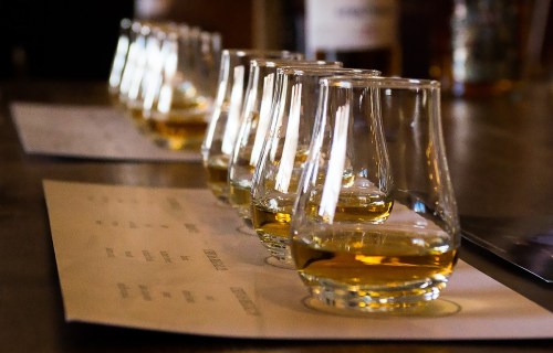 A row of whiskey tasting glasses filled with amber liquid, placed on a wooden table with tasting notes.