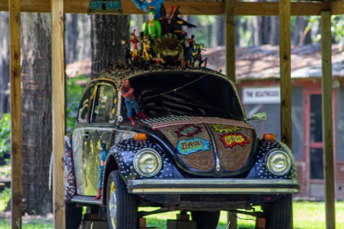 A colorful, decorated vintage car with action figures and comic-style graphics displayed under a wooden shelter.