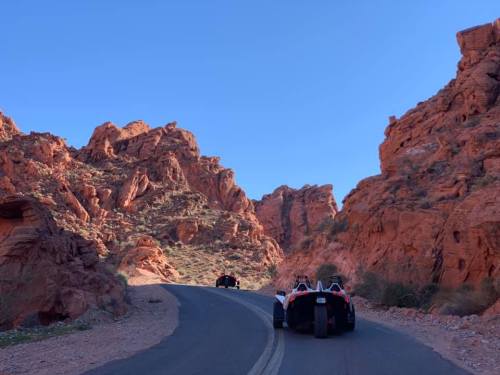 Two vehicles drive along a winding road through red rock formations under a clear blue sky.