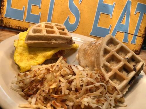 A plate with two waffle sandwiches filled with eggs and a side of crispy hash browns, with a sign that says "Let's Eat."