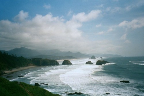A scenic coastal view with waves crashing on rocky shores, under a cloudy sky and distant mountains.