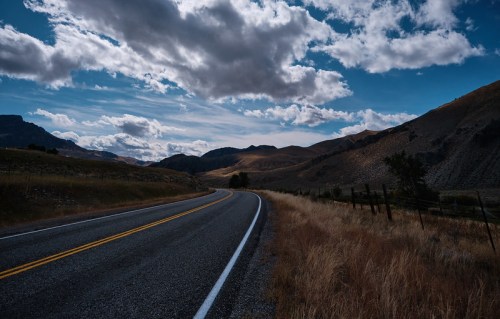 A winding road stretches through a mountainous landscape under a cloudy sky, with dry grass lining the edges.