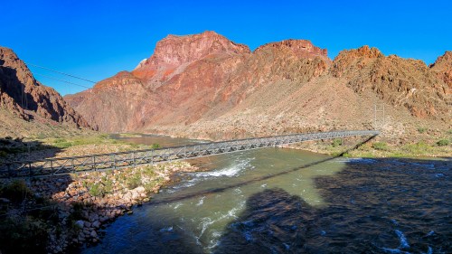 A metal bridge spans a river surrounded by rugged red rock formations under a clear blue sky.