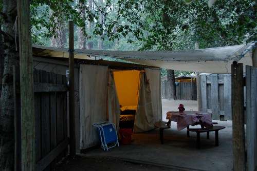A rustic campsite with a tent, picnic table, and string lights, surrounded by trees and wooden fences.