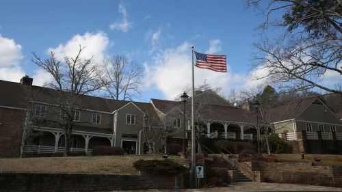 A building with a flagpole displaying the American flag, surrounded by trees and a blue sky with clouds.