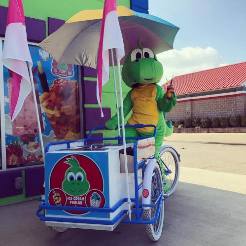 A green dinosaur mascot sits on an ice cream cart, holding a treat under a colorful umbrella.