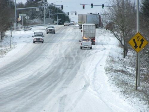 Snow-covered road with vehicles navigating, traffic lights in the background, and a warning sign for curves ahead.