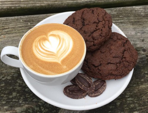 A cup of latte art with a heart design, accompanied by two chocolate cookies and a few chocolate pieces on a wooden table.