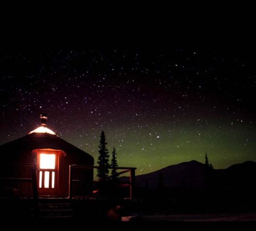 A cozy cabin illuminated at night, surrounded by trees under a starry sky with hints of the northern lights.