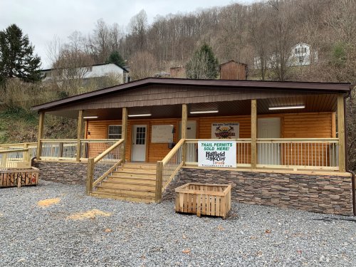 A wooden building with a porch, stone accents, and a sign for trail permits, surrounded by trees and hills.
