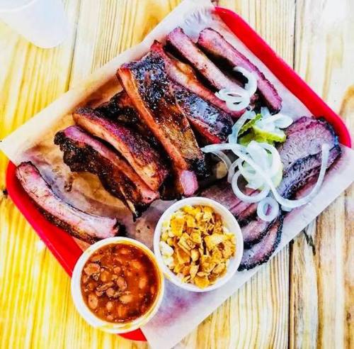 A red tray with smoked ribs, sliced onions, pickles, and sides of baked beans and coleslaw on a wooden table.