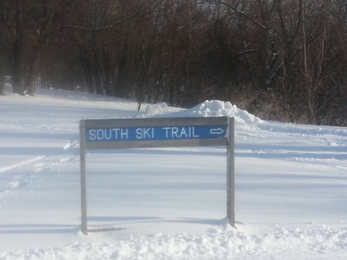 Sign for "South Ski Trail" pointing right, surrounded by a snowy landscape and trees in the background.
