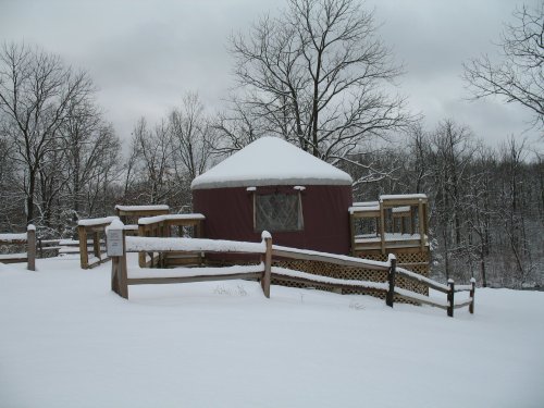 A snow-covered yurt surrounded by trees, with wooden railings and a cloudy sky overhead.