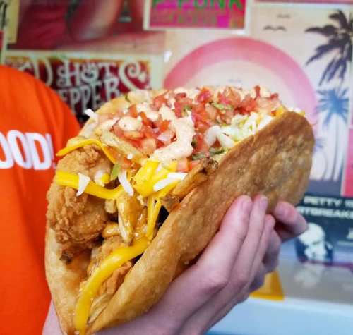 A person holding a large taco filled with fried chicken, cheese, lettuce, and diced tomatoes.