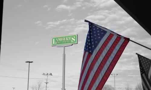 A large sign for Lambert's Cafe stands beside an American flag, with a cloudy sky in the background.