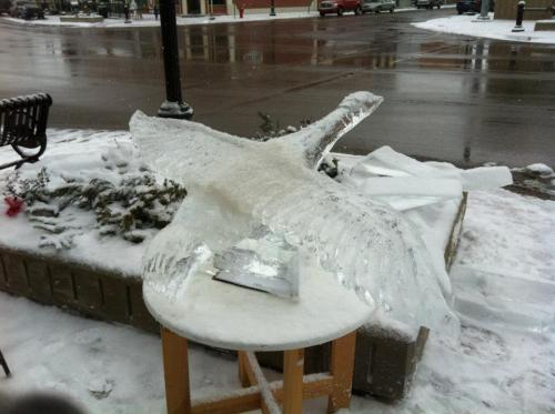 An ice sculpture of a bird in flight, displayed on a snowy street with a bench in the background.