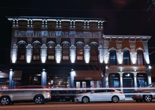 Historic brick building illuminated at night, featuring arched windows and a lively street scene with parked cars.