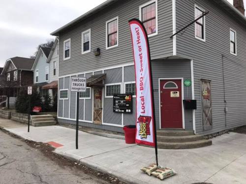 A gray building with a BBQ sign and a colorful flag, located on a street with nearby houses and a sidewalk.