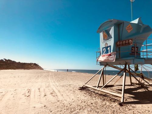 A lifeguard tower on a sandy beach under a clear blue sky, with gentle waves in the background.
