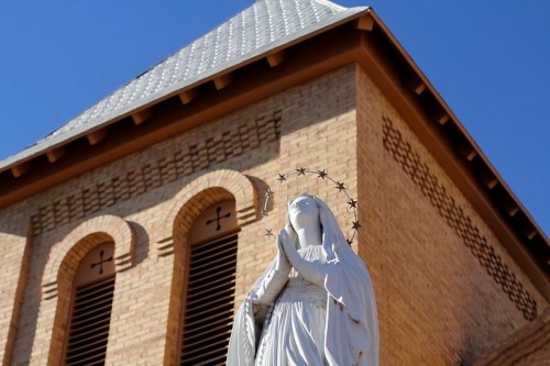 A white statue of a praying figure with a halo, set against a brick building and clear blue sky.