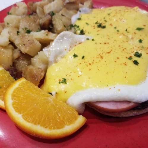 A plate of eggs Benedict with ham, hollandaise sauce, and seasoned potatoes, garnished with parsley and orange slices.