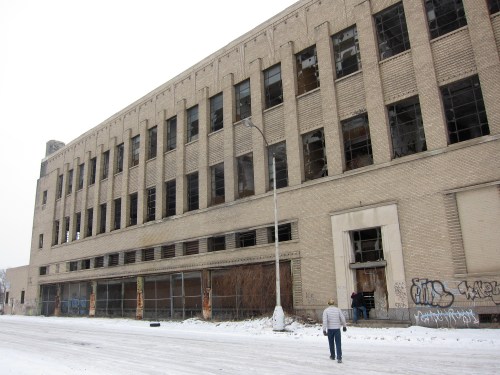A person walks towards a large, abandoned brick building with broken windows and graffiti, set in a snowy landscape.