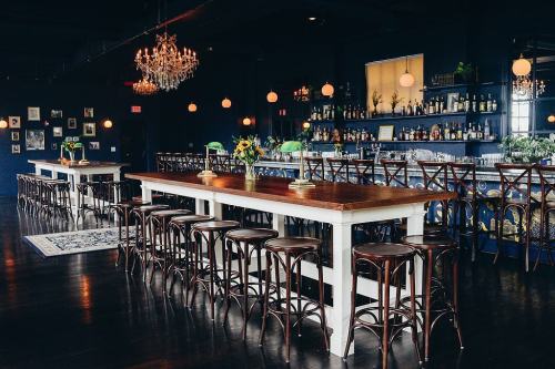 A stylish bar interior featuring a long wooden table, high stools, chandeliers, and a well-stocked bar in the background.