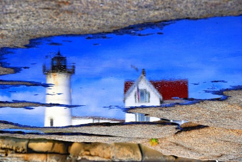 Reflection of a lighthouse and blue sky in a puddle on rocky terrain.