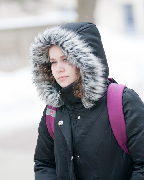 A young woman in a black parka with a fur-lined hood walks outdoors in winter, looking thoughtful. Snow is visible in the background.