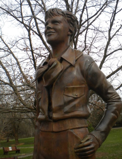 Bronze statue of a smiling aviator in a jacket, standing confidently in a park with bare trees in the background.