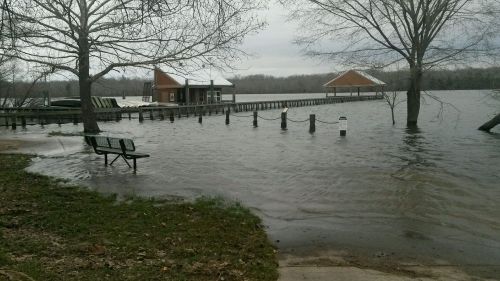 Flooded area with a bench, submerged dock, and buildings in the background under overcast skies.
