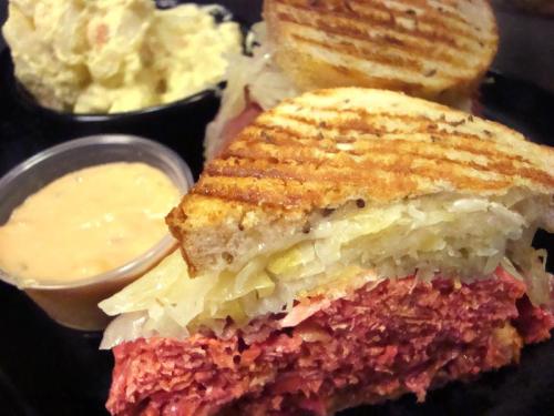 A close-up of a grilled sandwich with layers of meat and sauerkraut, served with a side of potato salad and sauce.
