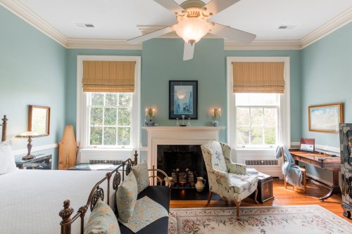 Cozy bedroom with blue walls, a fireplace, large windows, and vintage furniture, including a bed and armchair.