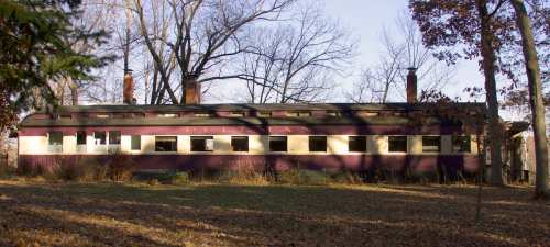 A vintage train car, the "Pelican," sits among trees in a wooded area, surrounded by fallen leaves.